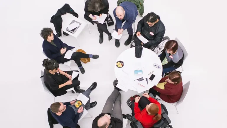 Contrasting the white floor, this photo shows an overhead view of a small group of people seated in a circle around a small white round table during a discussion or meeting. Several participants are holding papers or notebooks, and two people are using wheelchairs.