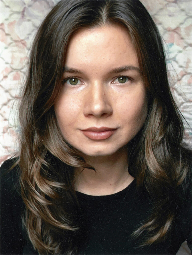 This is a portrait of Nadia Markiewicz. You can see her face centered, with shoulder-length brown hair and green eyes, looking directly at the camera with a neutral expression. She is wearing a black top, and the background is softly patterned in muted pastel tones.