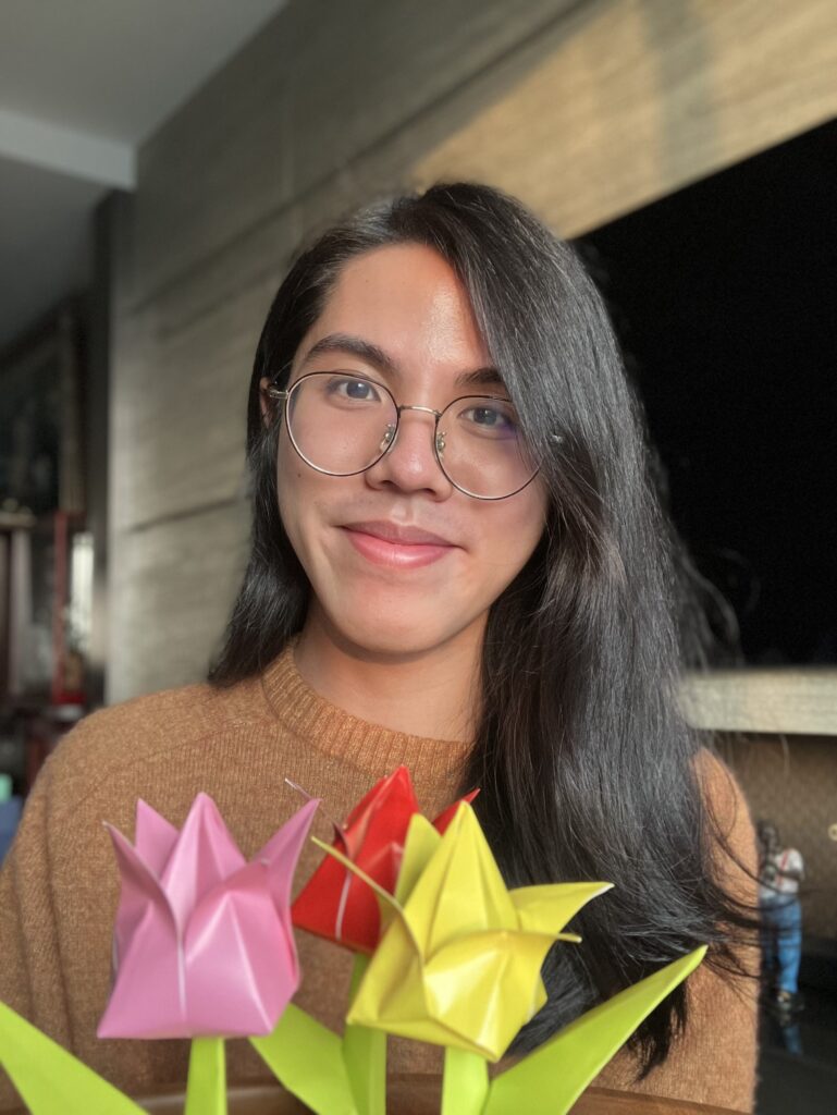 A tan Asian person with straight, shoulder-length black hair wearing round glasses and a burnt orange crew neck sweater. They are posing behind three origami tulips.