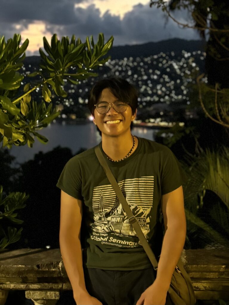 Leo is smiling, surrounded by greenery and is pictured in front of a lake.