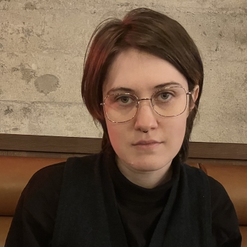 A young person with silver glasses and a serious expression stares into the camera. They have grey eyes, unevenly cut hair, and dark, layered clothing. A reddish glow illuminates one side of their face.