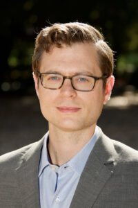 Professional headshot of Jeff Nagy, a white man with glasses standing outdoors in a gray suit. 
