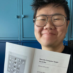 An image of Whit Pow, an Asian-American nonbinary trans scholar, with short cropped black hair and glasses, standing in front of a set of light and dark blue cabinets. They are holding a print copy of their article, "How the Computer Taught Us to See" in front of them at the bottom of the image. 