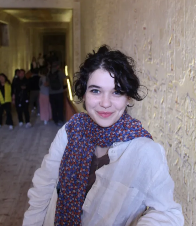 A photo of Phoebe More standing at the entrance to an Ancient Egyptian tomb in Luxor, Egypt. She is a white woman with blue eyes, dark, short hair pulled away from her face, and is wearing a white button down and a floral scarf around her neck. The stone walls behind her have hieroglyphic inscriptions and a group of tourists can be seen in the background. She is leaning on a railing facing the camera.