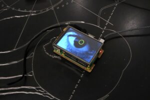 A photograph of an artwork taken from above. A digital image of an eye is on a screen on top of a circuit board with cables extending outside the frame. Thin metal wire creates loops and lines on a black surface.