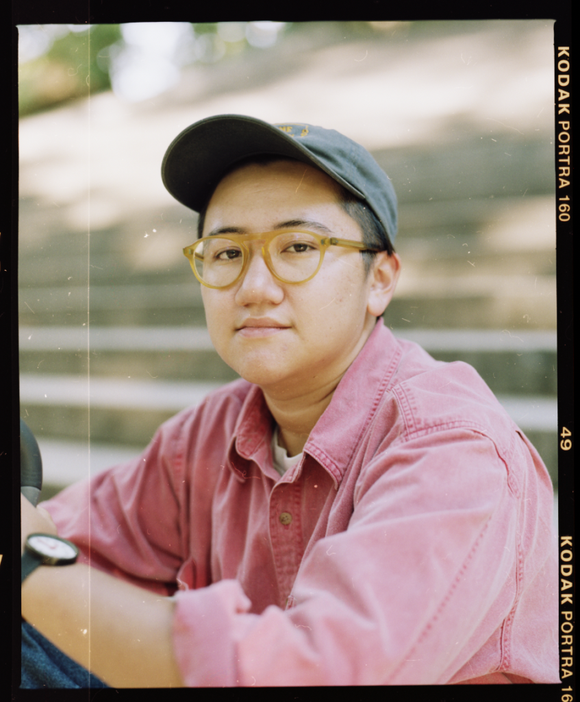 A photo of Emily Lim Rogers, wearing a light red collared shirt, glasses, and a baseball hat, seated in front of wide concrete steps in a blurry background. There are specks of light leaks on the photo suggesting it was printed and then photographed digitally. One edge is labeled, Kodak Porta 160, 49.